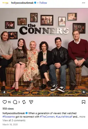 Michael Fishman sitting on a sofa alongside the ensemble cast of The Conners beneath framed family photos on the wall.