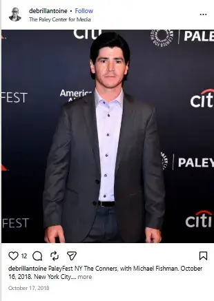 Michael Fishman standing in front of an event backdrop wearing a tailored charcoal grey suit over a light lavender dress shirt.