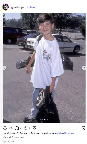 Young Michael Fishman smiling in a parking lot wearing an oversized white graphic t-shirt, jeans, and carrying gear bags.