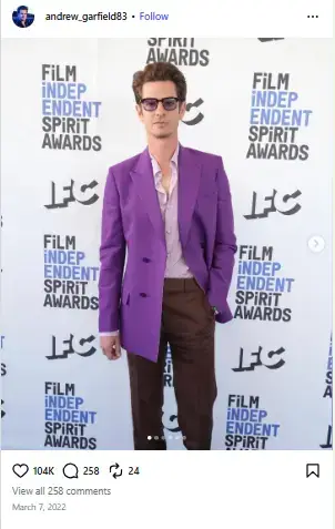 Andrew Garfield posing in a bright purple blazer, open-collar lavender shirt, sunglasses, and brown trousers on the Film Independent Spirit Awards red carpet.