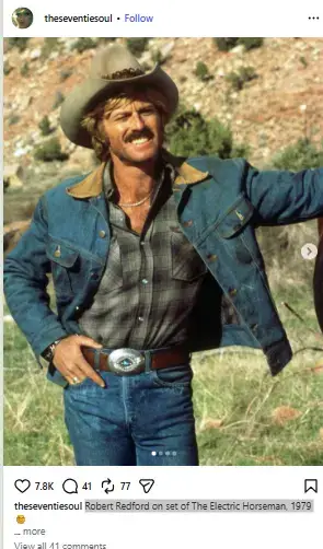 Robert Redford with a mustache wearing a cowboy hat, plaid shirt, denim jacket, and large belt buckle outdoors.