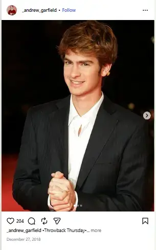 Young Andrew Garfield smiling with clasped hands in a black suit and open white collared shirt on a red carpet.