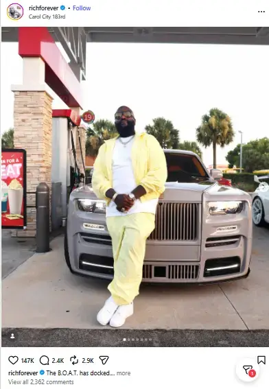 Rapper Rick Ross in a pale yellow tracksuit and sunglasses leaning against the front of a luxury grey Rolls-Royce at a gas station.