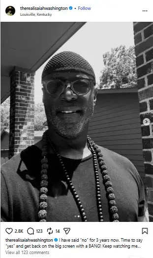 Black-and-white close-up of Isaiah Washington wearing glasses, a fitted beanie cap, and beaded necklaces while smiling outdoors.