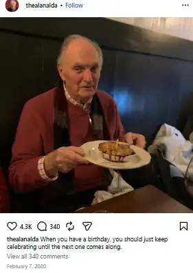 Alan Alda seated at a restaurant table holding up a plate with a small birthday dessert topped with a lit candle.