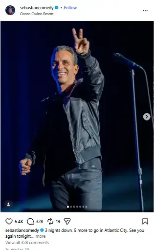 Sebastian Maniscalco smiling and flashing a peace sign on stage next to a microphone stand.