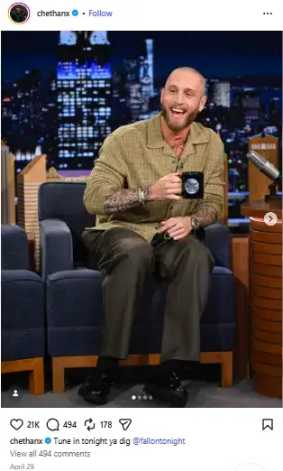 Chet Hanks smiling while seated as a guest on The Tonight Show Starring Jimmy Fallon, holding a show mug in an armchair.