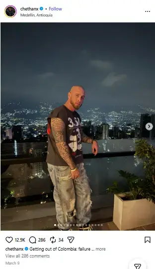 Chet Hanks showing off arm tattoos in a sleeveless graphic t-shirt and baggy jeans on a balcony overlooking Medellin at night.