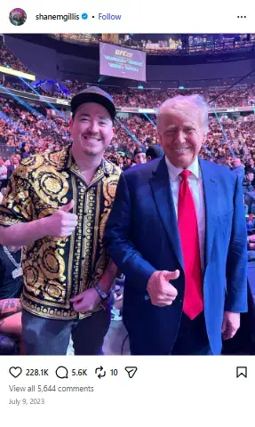 Shane Gillis in a patterned gold-and-black shirt smiling and giving a thumbs-up next to Donald Trump at a crowded UFC arena.