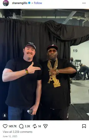 Shane Gillis pointing towards rapper Killer Mike wearing heavy gold chains and sunglasses backstage in front of a black curtain.