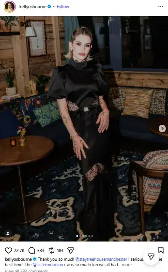 Kelly Osbourne posing in an elegant high-neck black satin gown with lace cutout detailing inside a stylish lounge.