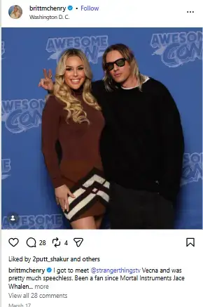 Britt McHenry posing side by side with actor Jamie Campbell Bower in sunglasses in front of an Awesome Con backdrop.