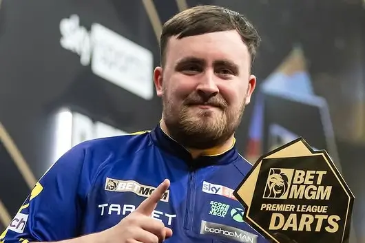 Luke Littler smiling and pointing while holding his Premier League Darts trophy.