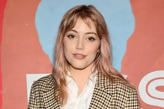 Grace Van Dien, smiles at the camera, wearing a plaid blazer over a white shirt.