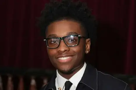 Miles Caton, wearing glasses, a tie, and a dark jacket.