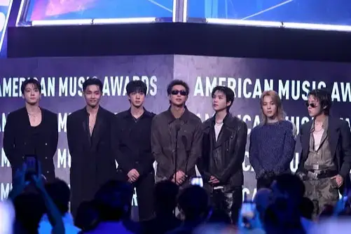 The seven members of BTS stand on stage together in front of an "American Music Awards"