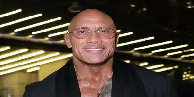 Smiling and bald Dwayne "The Rock" Johnson wearing glasses, a black suit.